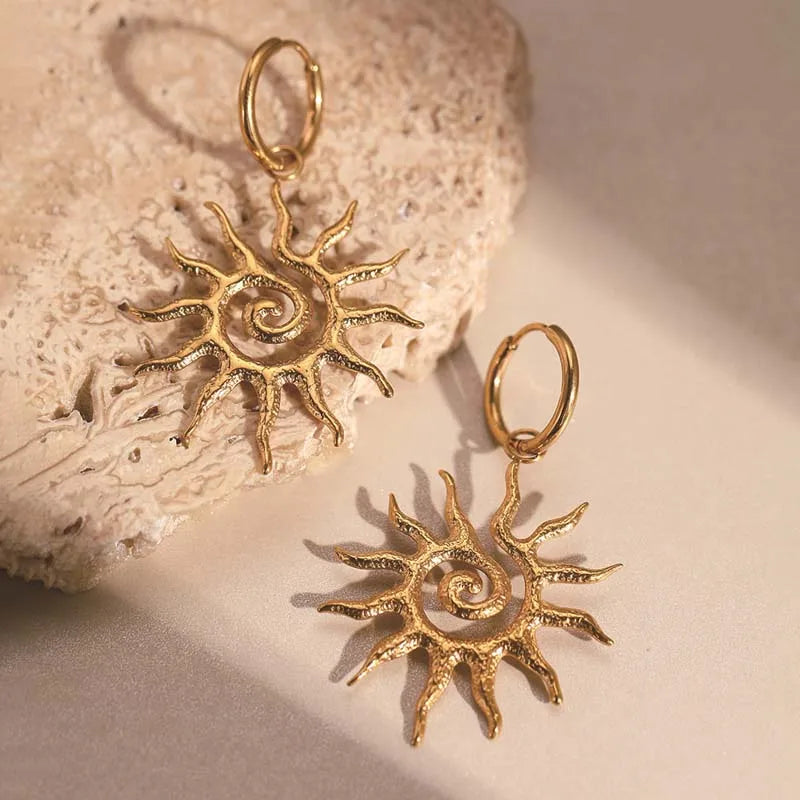 Gold sun-shaped earrings on a textured beige surface. Bishop X 