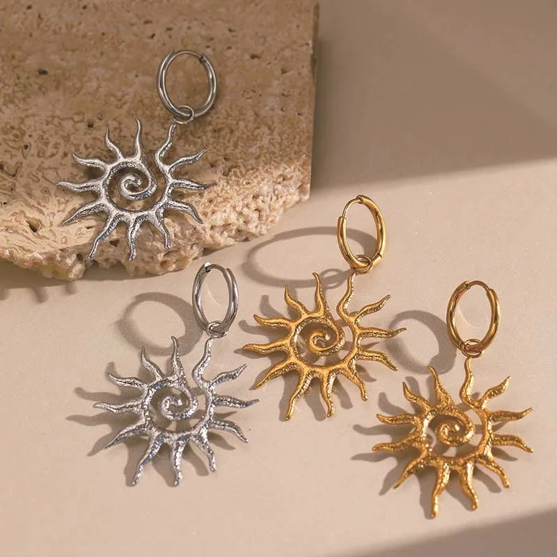 Sun-shaped earrings in silver and gold on a beige surface with a stone background. Bishop x